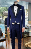 A man wearing a blue three-piece tuxedo with a jacket, waistcoat, and trousers, featuring a black bow tie and pocket square.